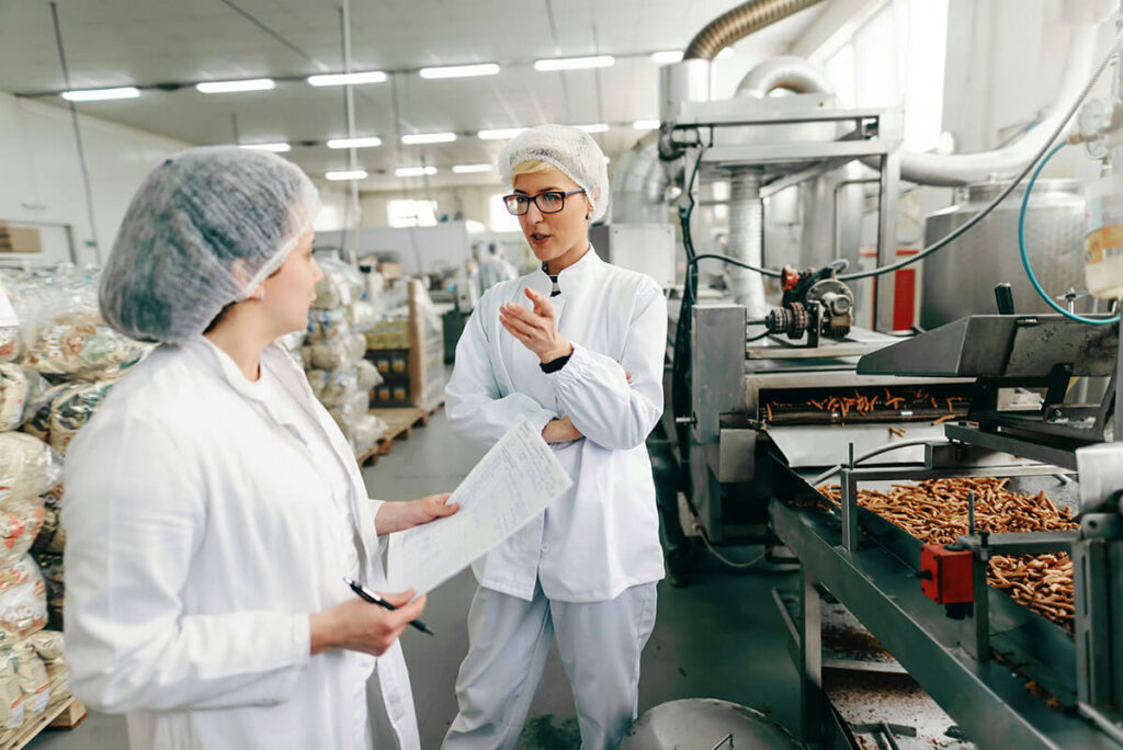 Two women talking while standing next to an assembly line of food.